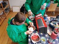 Zwei Kinder, die grüne Malkittel tragen, sind dabei, ein Objekt rot und weiß zu bemalen. Um sie herum sind verschiedene Farben, Pinsel und Becher mit Flüssigkeit zu sehen.